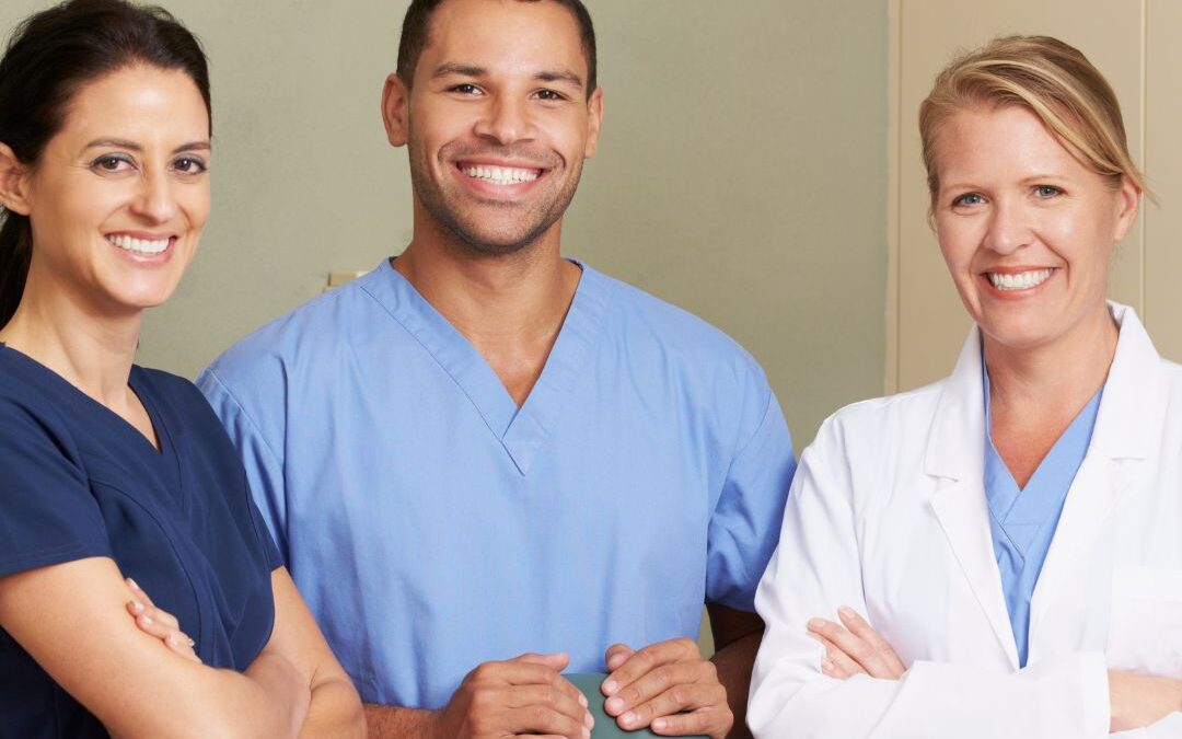 dental team of three standing and smiling wearing scrubs - demale with crossed arms, male with hands on chair, female with crossed arms