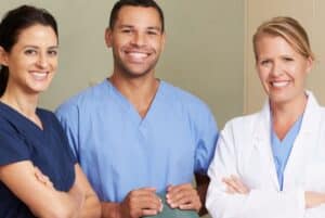 dental team of three standing and smiling wearing scrubs - demale with crossed arms, male with hands on chair, female with crossed arms