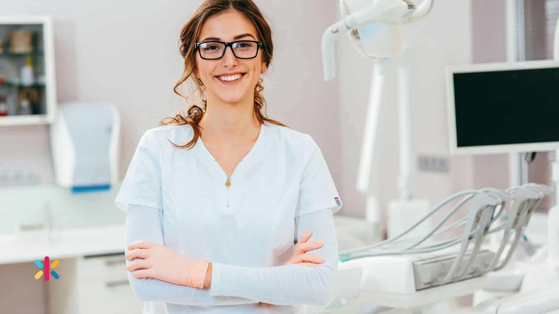 Female dental professional wearing glasses smiling confidently in a dental clinic with tools and monitor